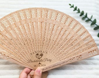 May include: A light brown wooden hand fan with intricate cut-out designs. The fan is inscribed with "SANDRA'S Bachelorette Party" and the date "08.14.2026". The fan also has the phrase "Thanks for celebrating with me!"