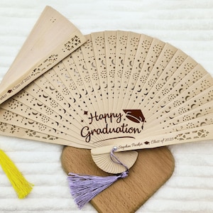 May include: A wooden hand fan with a cutout design of stars and moons. The fan is personalized with the text "Happy Graduation!" and the name "Stephen Parker" with the year "Class of 2023".