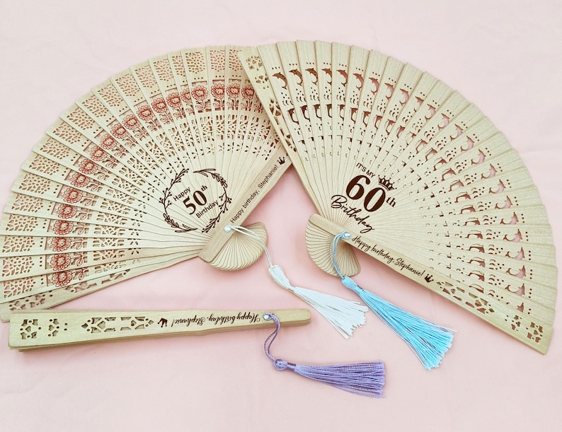 Op de afbeelding: Twee houten vouwvaandels met decoratieve uitsnijdingen en kwastjes. De grotere waaier heeft de tekst "It's my 60th Birthday" en "Happy birthday, Stephanie!" De kleinere waaier heeft de tekst "Happy 50th Birthday" en "Happy birthday, Stephanie!"
