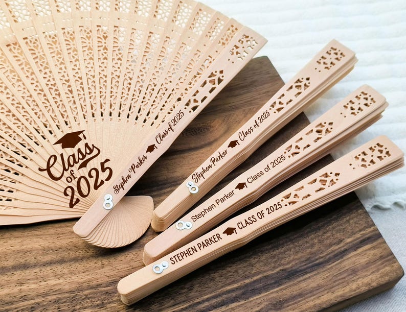 May include: A collection of personalized wooden fans with laser-cut designs. One fan is fully open, displaying "Class of 2025" and the name "Stephen Parker." Three closed fans are also visible, each engraved with the same text and name. The fans are light brown.