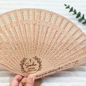 May include: A wooden hand fan with a delicate, intricate design. It features a personalized message with a cross, floral wreath, and the text "Sandra Baptized June 27, 2025". The fan also includes the text "Thank you for celebrating with us."