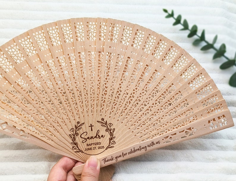 May include: A wooden hand fan with a delicate, intricate design. It features a personalized message with a cross, floral wreath, and the text "Sandra Baptized June 27, 2025". The fan also includes the text "Thank you for celebrating with us."