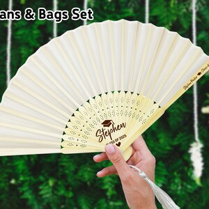 May include: White hand fan with a brown wooden handle and a white fabric panel. The fan has a decorative design with a heart and the text "Stephen Class of 2023". The back of the fan has the text "Stephen Parker Class of 2023".
