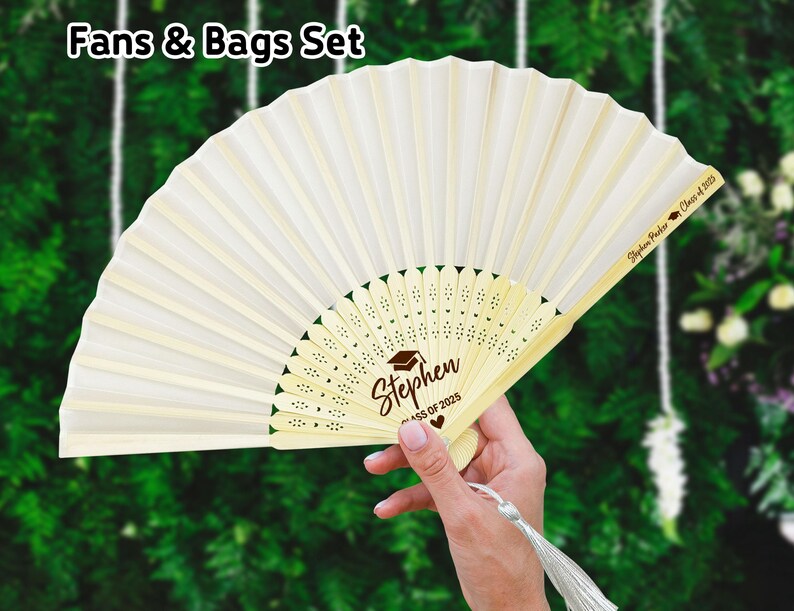 May include: White hand fan with a brown wooden handle and a white fabric panel. The fan has a decorative design with a heart and the text "Stephen Class of 2023". The back of the fan has the text "Stephen Parker Class of 2023".