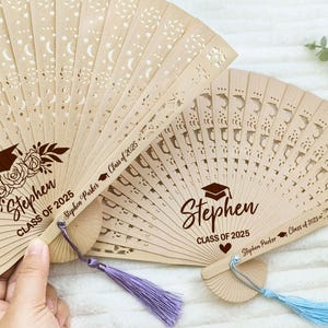 May include: Two wooden hand fans with a graduation theme. The fans are decorated with a star and moon pattern and have the text "Stephen Class of 2025" printed on them.