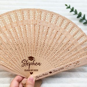 May include: A light brown wooden fan with intricate cut-out designs. The fan is personalized with the name "Stephen", a graduation cap, and the text "CLASS OF 2025". The fan is held open, displaying its full shape.