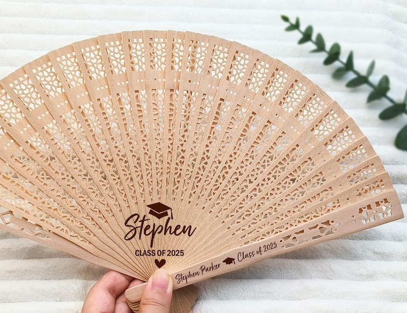 May include: A light brown wooden fan with intricate cut-out designs. The fan is personalized with the name "Stephen", a graduation cap, and the text "CLASS OF 2025". The fan is held open, displaying its full shape.