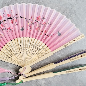 Puede incluir: Tres abanicos plegables de color rosa y verde con un diseño de flor de cerezo. Los abanicos tienen un mango de madera y están decorados con el texto "Stephen Parker Class of 2025".