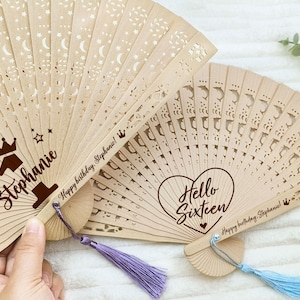 Op de afbeelding: Twee houten handventilatoren met decoratieve uitsparingen en gepersonaliseerde berichten. De eerste ventilator heeft een kroon en het nummer &eacute;&eacute;n met de naam Stephanie en het bericht "Happy birthday Stephanie!" De tweede ventilator heeft een hart met de tekst "Hello Sixteen" en het bericht "Happy birthday Stephanie!"