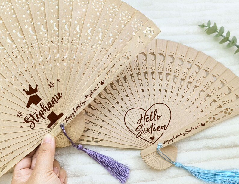 Op de afbeelding: Twee houten handventilatoren met decoratieve uitsparingen en gepersonaliseerde berichten. De eerste ventilator heeft een kroon en het nummer &eacute;&eacute;n met de naam Stephanie en het bericht "Happy birthday Stephanie!" De tweede ventilator heeft een hart met de tekst "Hello Sixteen" en het bericht "Happy birthday Stephanie!"