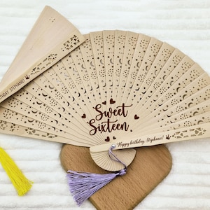 Op de afbeelding: Een houten handwaaier met een kantontwerp en de woorden "Sweet Sixteen" en "Happy Birthday Stephanie!" De waaier heeft een gele kwast en een paarse kwast.