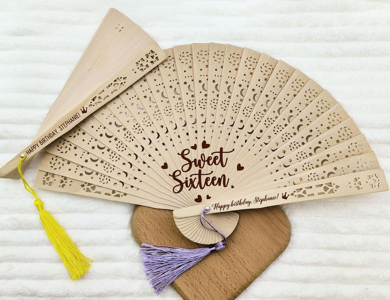 Op de afbeelding: Een houten handwaaier met een kantontwerp en de woorden "Sweet Sixteen" en "Happy Birthday Stephanie!" De waaier heeft een gele kwast en een paarse kwast.