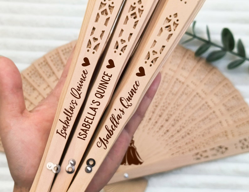 Puede incluir: Tres abanicos de mano de madera con un dise&ntilde;o decorativo de corte. Cada abanico tiene un coraz&oacute;n y el texto "Isabella's Quince" impreso en &eacute;l.
