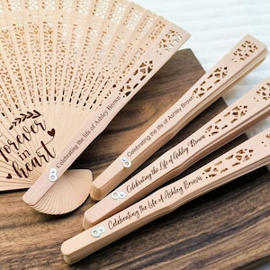 May include: A set of four wooden hand fans with a delicate, intricate design. The fans are engraved with the text "Forever in heart" and "Celebrating the life of Ashley Brown".