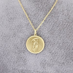 Op de afbeelding: Een gouden ketting met een ronde hanger met een gedetailleerde gravure van de Grim Reaper die een zeis vasthoudt.