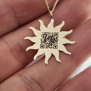 May include: A gold-colored sun pendant necklace. The sunburst design features multiple rays and a QR code in the center. The necklace is displayed on a hand.