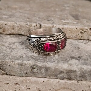 Puede incluir: Un anillo de plata con un intrincado diseño floral y tres piedras preciosas rojas. El anillo está colocado sobre una superficie de piedra texturizada.