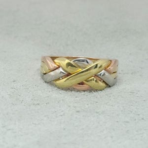Può includere: Un anello puzzle composto da quattro fasce intrecciate in metallo lucido. Il design presenta una combinazione di fasce in oro giallo, oro rosa e color argento, intrecciate insieme per formare un'unica struttura circolare coesa.