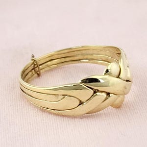 May include: A gold puzzle ring composed of four interlocking bands that weave together to form a single cohesive unit. The polished metal surface reflects light, highlighting the intricate, overlapping design of the connected bands.