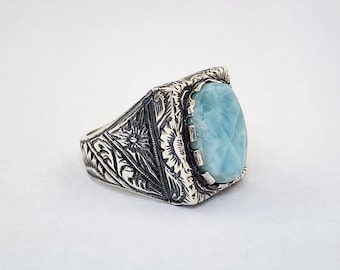 May include: A silver ring with an ornate design and a light blue gemstone. The ring has a square setting with a detailed floral pattern.