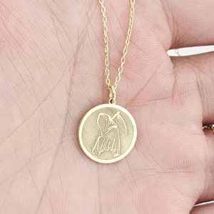 Op de afbeelding: Een gouden ketting met een ronde hanger met een gedetailleerd silhouet van de Grim Reaper die een zeis vasthoudt.