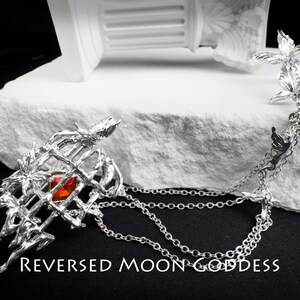 K&ouml;nnte beinhalten: Silberne Halskette mit einem K&auml;fig-Anh&auml;nger mit einem roten Edelstein und einer Kette mit mehreren Vogelanh&auml;ngern. Der Text "REVERSED MOON GODDESS" ist am unteren Rand des Bildes sichtbar.