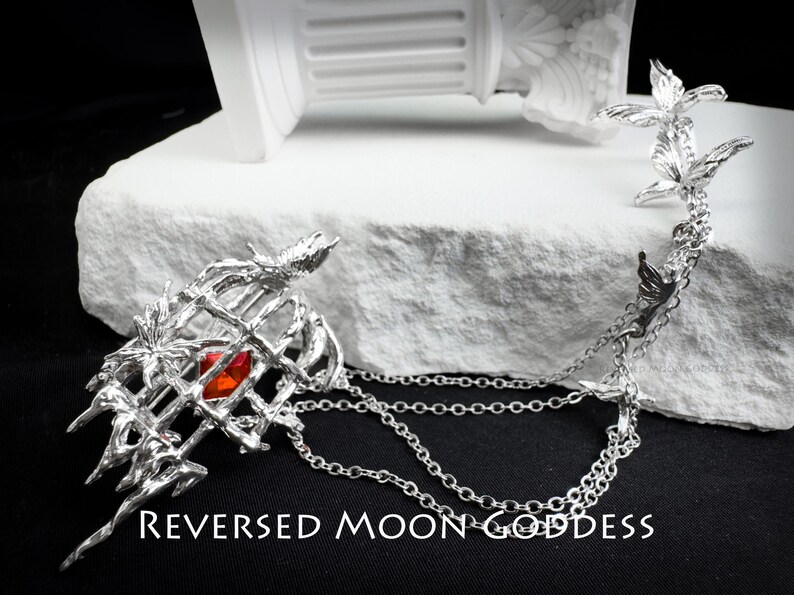 K&ouml;nnte beinhalten: Silberne Halskette mit einem K&auml;fig-Anh&auml;nger mit einem roten Edelstein und einer Kette mit mehreren Vogelanh&auml;ngern. Der Text "REVERSED MOON GODDESS" ist am unteren Rand des Bildes sichtbar.