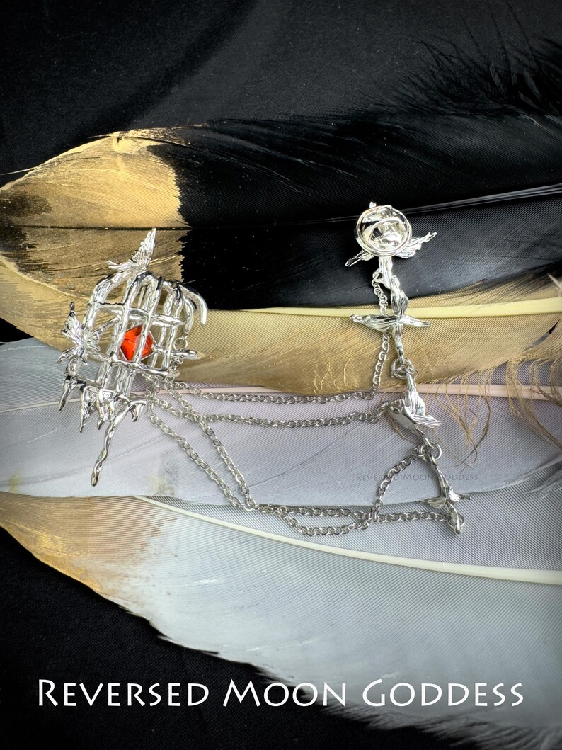 K&ouml;nnte beinhalten: Zwei silberne Schmuckst&uuml;cke, die durch eine Kette verbunden sind. Das St&uuml;ck links ist ein K&auml;fig mit einem roten Edelstein darin. Das St&uuml;ck rechts ist eine Rose mit einem Vogel, der oben drauf sitzt. Der Schmuck befindet sich auf einem wei&szlig;en und goldenen Federhintergrund. Der Text "REVERSED MOON GODDESS" befindet sich am unteren Rand des Bildes.
