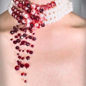 Puede incluir: Un collar de varias capas con cuentas blancas y rojas. El collar tiene un dise&ntilde;o en cascada con cuentas de cristal rojas que cuelgan de la parte inferior. El texto "REVERSED MOON GODDESS" est&aacute; impreso en la parte inferior de la imagen.