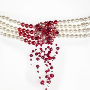 Puede incluir: Un collar de cuentas rojas y blancas con un racimo de cuentas rojas en el centro y una hebra de cuentas rojas colgantes. El collar se llama "REVERSED MOON GODDESS".