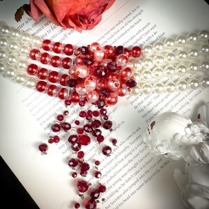 Puede incluir: Un collar de cuentas rojas y blancas en forma de coraz&oacute;n, esparcido sobre un libro con el texto "REVERSED MOON GODDESS".