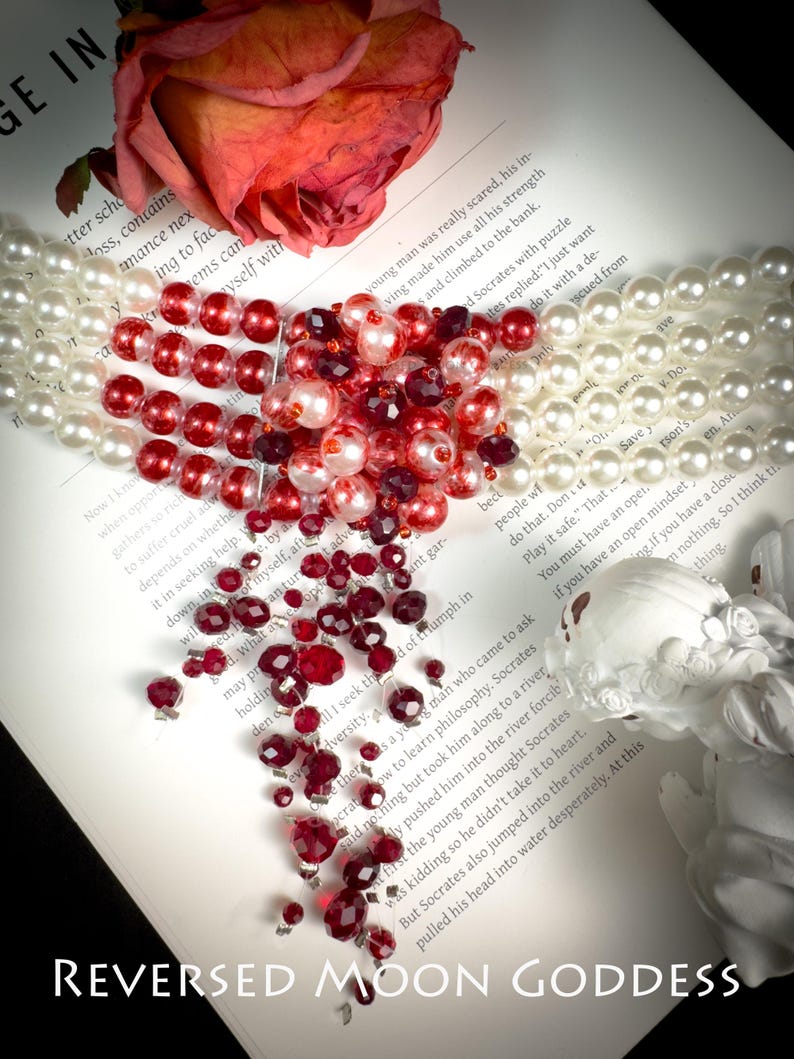 Puede incluir: Un collar de cuentas rojas y blancas en forma de coraz&oacute;n, esparcido sobre un libro con el texto "REVERSED MOON GODDESS".