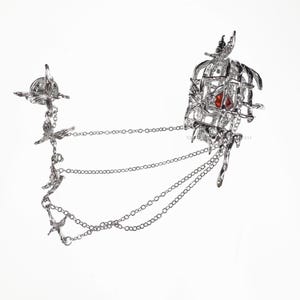 K&ouml;nnte beinhalten: Silberner Schmuck mit einem Vogelk&auml;fig mit einem roten Edelstein darin und mehreren Ketten mit Vogelanh&auml;ngern. Der Text "REVERSED MOON GODDESS" befindet sich am unteren Rand des Bildes.