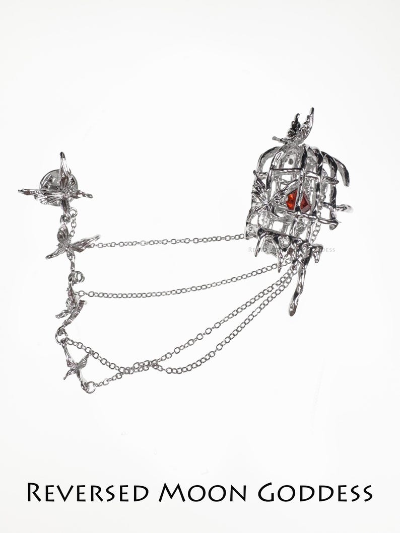K&ouml;nnte beinhalten: Silberner Schmuck mit einem Vogelk&auml;fig mit einem roten Edelstein darin und mehreren Ketten mit Vogelanh&auml;ngern. Der Text "REVERSED MOON GODDESS" befindet sich am unteren Rand des Bildes.