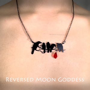 May include: A black necklace with a pendant featuring five black birds perched on a branch, with a red gemstone dangling below. The necklace is on a person's chest, and the text "REVERSED MOON GODDESS" is visible below the pendant.