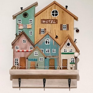 Może przedstawiać: Drewniany, ścienny wieszak na klucze w kształcie szeregu kolorowych domków. Domki mają okna, drzwi i dachy. Środkowy domek ma tabliczkę z napisem "HOTEL".  Wieszak ma trzy haczyki na dole.