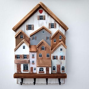 Può includere: Un portachiavi in legno a forma di villaggio. Le case sono dipinte in varie tonalità di marrone, bianco e grigio, con piccole finestre e persiane. Un cuore rosso decora la parte superiore. Il portachiavi ha un ripiano con quattro ganci.