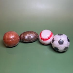 May include: Four sports-themed bath bombs. A brown football, a brown football, a white baseball with red stitching, and a soccer ball with black pentagons and white hexagons. All are wrapped in clear plastic.