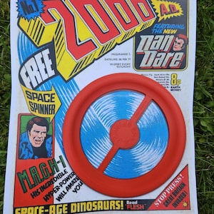 Puede incluir: Una portada de cómic vintage "2000 A.D." con un juguete giratorio espacial rojo y azul. El cómic presenta el texto "Dan Dare" y "Space-Age Dinosaurs!" y la fecha "26 FEB 77".