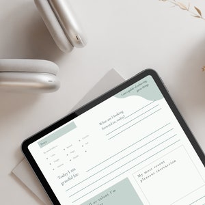 May include: A digital tablet with a white background and a light blue and green design. The tablet is displaying a daily planner with sections for date, mood, gratitude, goals, and a recent pleasant interaction. The text on the tablet reads "Date", "Today I am grateful for", "What am I looking forward to today?", "I am capable of achieving great things", and "My most recent pleasant interaction".