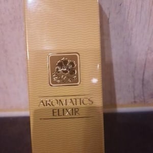 Może przedstawiać: Pudełko perfum w sprayu Aromatics Elixir. Pudełko jest w jasnobeżowym kolorze z falistym wzorem. Tekst "Aromatics Elixir" jest złotymi literami. Złoty kwadrat z kwiatowym wzorem znajduje się z przodu. Zawiera 45 ml.
