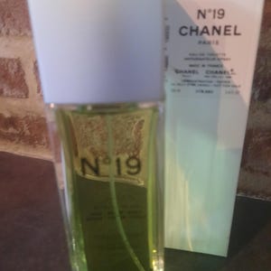 Pu&ograve; includere: Un flacone rettangolare in vetro di Chanel N&deg;19 eau de toilette con liquido verde chiaro, dotato di un tappo bianco e testo nero sulla parte anteriore. Accanto si trova una scatola bianca alta con la scritta N&deg;19 CHANEL PARIS stampata in nero.