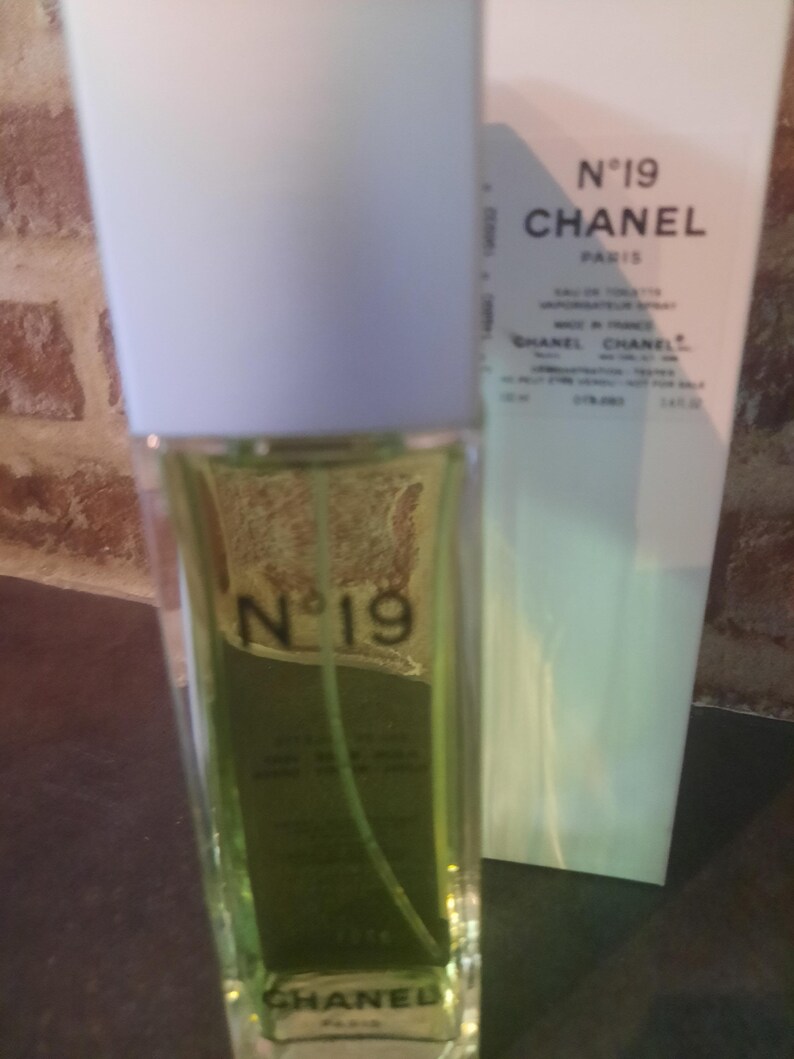 Pu&ograve; includere: Un flacone rettangolare in vetro di Chanel N&deg;19 eau de toilette con liquido verde chiaro, dotato di un tappo bianco e testo nero sulla parte anteriore. Accanto si trova una scatola bianca alta con la scritta N&deg;19 CHANEL PARIS stampata in nero.