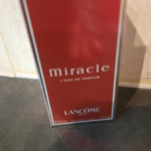 Puede incluir: Una caja roja con un borde plateado y la palabra "miracle" impresa en blanco. La caja también tiene las palabras "Lancôme Paris" impresas en blanco.