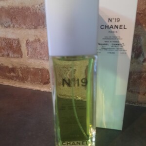Pu&ograve; includere: Un flacone rettangolare in vetro di Chanel N&deg;19 Eau de Toilette con liquido verde chiaro, tappo bianco e testo nero sul davanti. Accanto si trova una scatola rettangolare bianca alta che riporta il testo N&deg;19 CHANEL PARIS e altri dettagli.