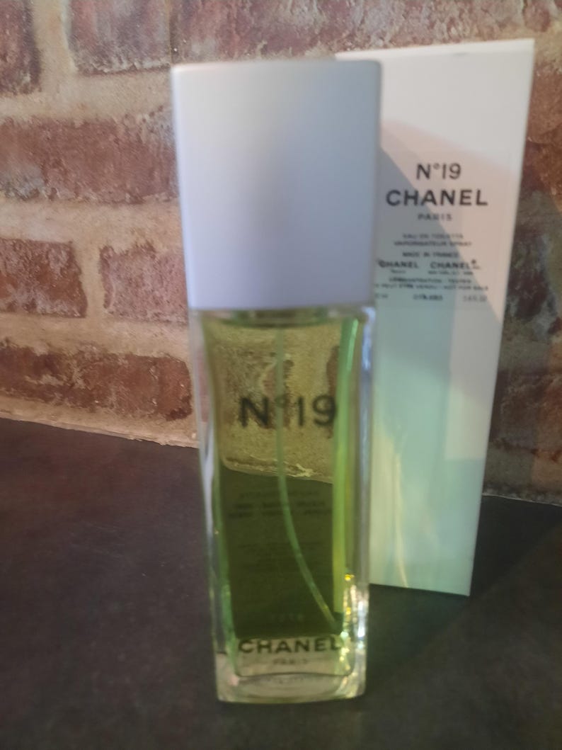 Pu&ograve; includere: Un flacone rettangolare in vetro di Chanel N&deg;19 Eau de Toilette con liquido verde chiaro, tappo bianco e testo nero sul davanti. Accanto si trova una scatola rettangolare bianca alta che riporta il testo N&deg;19 CHANEL PARIS e altri dettagli.