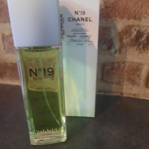 Pu&ograve; includere: Un flacone rettangolare in vetro di Chanel N&deg;19 eau de toilette con liquido verde chiaro e tappo bianco, posizionato davanti alla sua scatola di cartone bianca abbinata. Sia il flacone che la scatola riportano il marchio Chanel N&deg;19 in nero.
