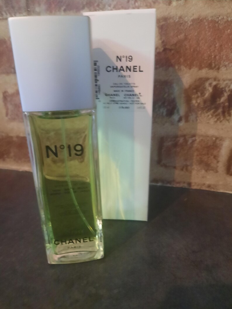 Pu&ograve; includere: Un flacone rettangolare in vetro di Chanel N&deg;19 eau de toilette con liquido verde chiaro e tappo bianco, posizionato davanti alla sua scatola di cartone bianca abbinata. Sia il flacone che la scatola riportano il marchio Chanel N&deg;19 in nero.