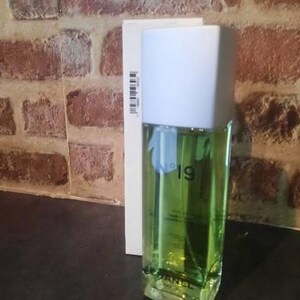 Pu&ograve; includere: Un flacone di profumo rettangolare in vetro riempito di liquido verde chiaro, dotato di un tappo bianco e della scritta N&deg;19 CHANEL stampata sul davanti. Il flacone &egrave; posizionato davanti alla sua scatola di cartone bianco, alta e stretta.