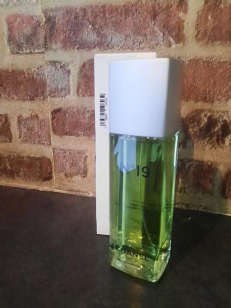 Pu&ograve; includere: Un flacone di profumo rettangolare in vetro riempito di liquido verde chiaro, dotato di un tappo bianco e della scritta N&deg;19 CHANEL stampata sul davanti. Il flacone &egrave; posizionato davanti alla sua scatola di cartone bianco, alta e stretta.